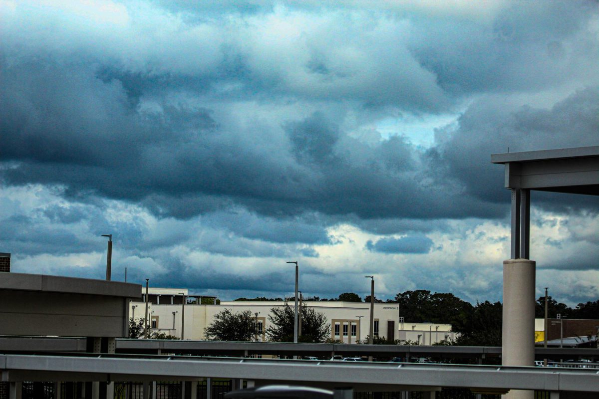 This photo shows a view of the sky with dark blue clouds, representing a storm. This was taken from above in building 4, in the background you can see the buildings of the other institution Cypress Creek Middle School which is surrounded by a forest in the back. 
Esta foto muestra una vista del cielo con nubes de color azul oscuro, representando una tormenta. Esta fue tomada desde arriba en el edificio 4, al fondo se pueden ver los edificios de la otra institución Cypress Creek Middle School que está rodeada por un bosque en la parte de atrás.